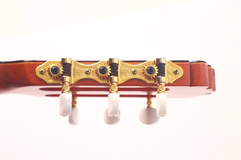 Nahaufnahme der goldenen Stimmwirbel und Zahnräder an der Kopfplatte einer Juan Miguel Gonzalez 2006 Klassische Gitarre mit weißen Stimmschlüsseln und polierter Holzoberfläche vor einem hellen Hintergrund. Nahaufnahme der goldenen Stimmwirbel und Zahnräder an der Kopfplatte einer Juan Miguel Gonzalez 2006 Klassische Gitarre mit weißen Stimmschlüsseln und polierter Holzoberfläche vor einem hellen Hintergrund.