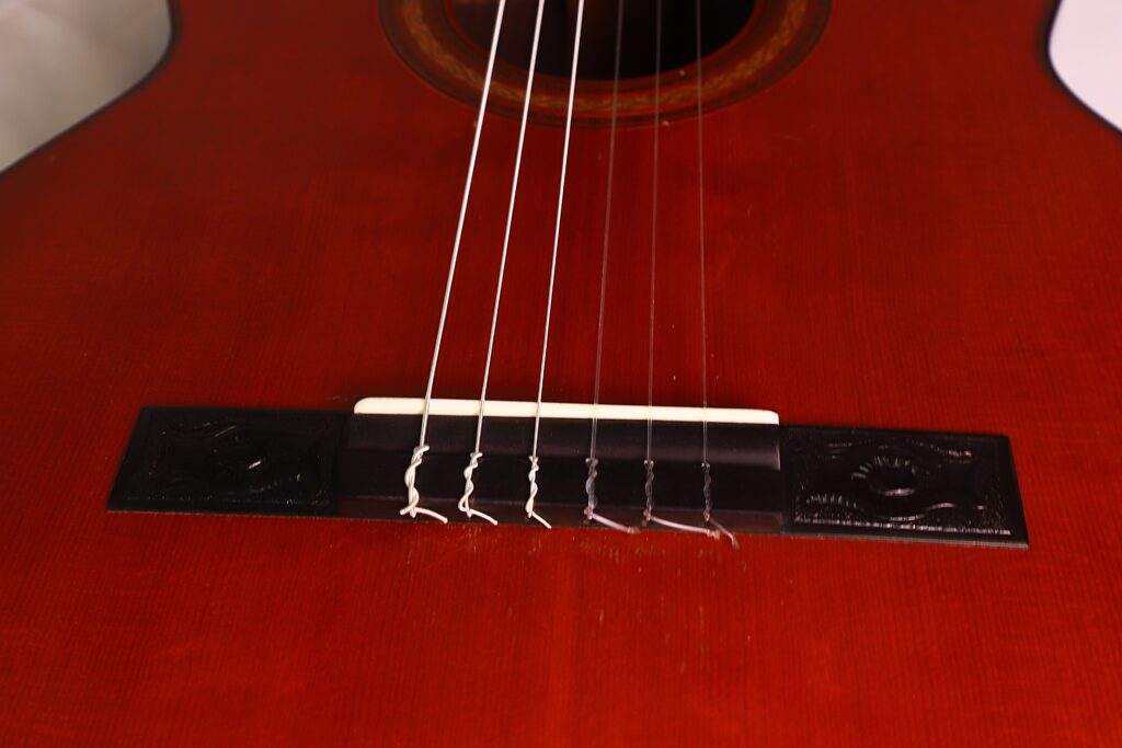Nahaufnahme des Stegs und der Knoten der Nylonsaiten an der „Klassische Gitarre“ von Joachim Schneider aus dem Jahr 1977, die die rötliche Holzoberfläche besonders gut zur Geltung bringt. Nahaufnahme des Stegs und der Knoten der Nylonsaiten an der „Klassische Gitarre“ von Joachim Schneider aus dem Jahr 1977, die die rötliche Holzoberfläche besonders gut zur Geltung bringt.