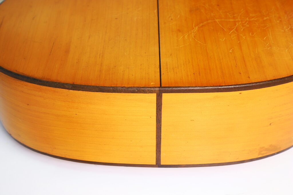Nahaufnahme des Bodens und der Seite einer Jose Ramirez I 1921 Klassische Gitarre, die die Holzmaserung, die dunklen Zierleisten und die leichten Oberflächenkratzer hervorhebt. Der Fokus liegt auf der Fuge, an der die Bodenplatten der Gitarre aufeinandertreffen. Nahaufnahme des Bodens und der Seite einer Jose Ramirez I 1921 Klassische Gitarre, die die Holzmaserung, die dunklen Zierleisten und die leichten Oberflächenkratzer hervorhebt. Der Fokus liegt auf der Fuge, an der die Bodenplatten der Gitarre aufeinandertreffen.