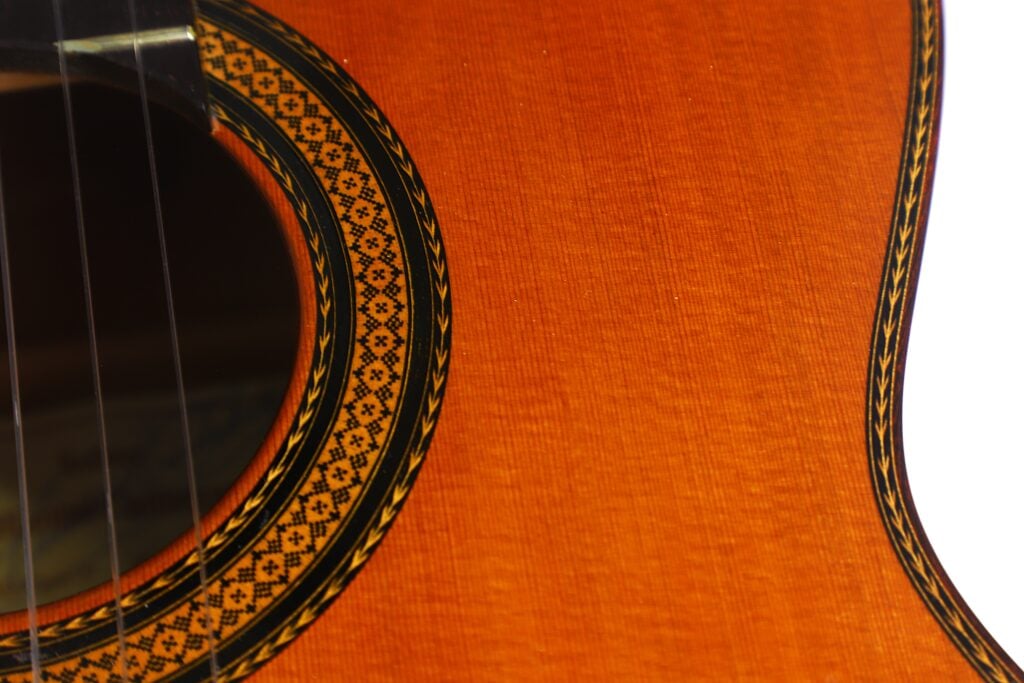 Nahaufnahme des Korpus der „Doble Tapa Klassische Gitarre“ von Manuel Contreras aus dem Jahr 1981, die die detailreiche Zierrosette um das Schallloch und die glatte orange-braune Holzmaserung zeigt, wobei links teilweise die Saiten zu sehen sind. Nahaufnahme des Korpus der „Doble Tapa Klassische Gitarre“ von Manuel Contreras aus dem Jahr 1981, die die detailreiche Zierrosette um das Schallloch und die glatte orange-braune Holzmaserung zeigt, wobei links teilweise die Saiten zu sehen sind.