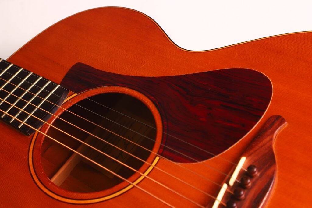 Nahaufnahme der Lowden S-5 Akustikgitarre, die die Saiten, das Schallloch, das hölzerne Schlagbrett und einen Teil des Stegs auf einem sattbraunen Holzkorpus zeigt. Nahaufnahme der Lowden S-5 Akustikgitarre, die die Saiten, das Schallloch, das hölzerne Schlagbrett und einen Teil des Stegs auf einem sattbraunen Holzkorpus zeigt.