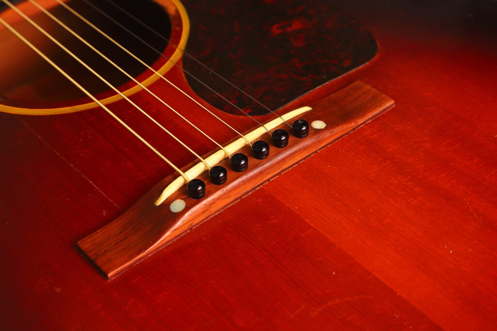 Nahaufnahme der Gibson J-45 Banner 1944 Akustik Gitarre mit sechs Saiten, Holzsteg und einem Teil des dunklen Schlagbretts auf dem rötlich-braunen Korpus. Nahaufnahme der Gibson J-45 Banner 1944 Akustik Gitarre mit sechs Saiten, Holzsteg und einem Teil des dunklen Schlagbretts auf dem rötlich-braunen Korpus.