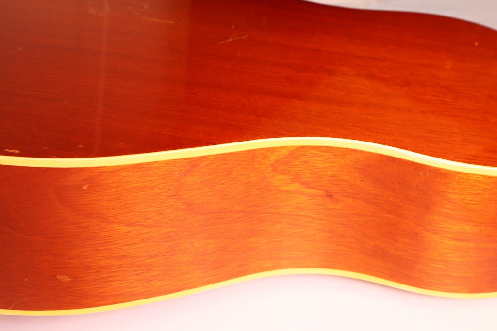 Nahaufnahme der Seite der Gibson J-50 Akustikgitarre aus dem Jahr 1952, die die glatte, polierte Holzoberfläche und die zarten, hellen Kantenverzierungen zeigt. Nahaufnahme der Seite der Gibson J-50 Akustikgitarre aus dem Jahr 1952, die die glatte, polierte Holzoberfläche und die zarten, hellen Kantenverzierungen zeigt.