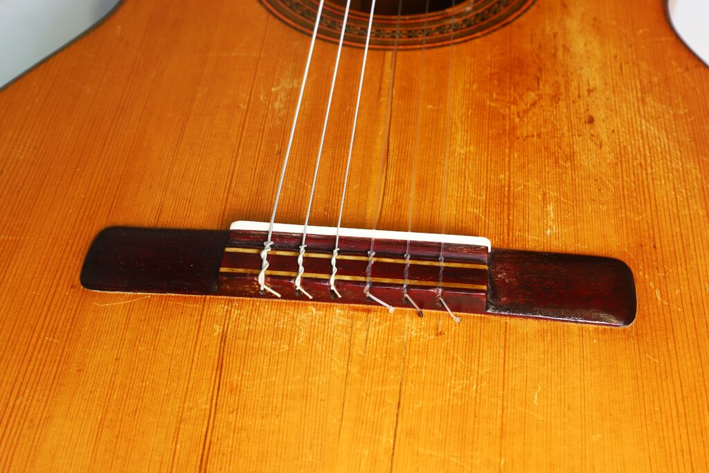 Nahaufnahme des Stegs und der Saiten einer Jose Ramirez I 1921 Klassische Gitarre, die die Maserung des Holzes und die sichtbaren Kratzer am Korpus hervorhebt. Nahaufnahme des Stegs und der Saiten einer Jose Ramirez I 1921 Klassische Gitarre, die die Maserung des Holzes und die sichtbaren Kratzer am Korpus hervorhebt.