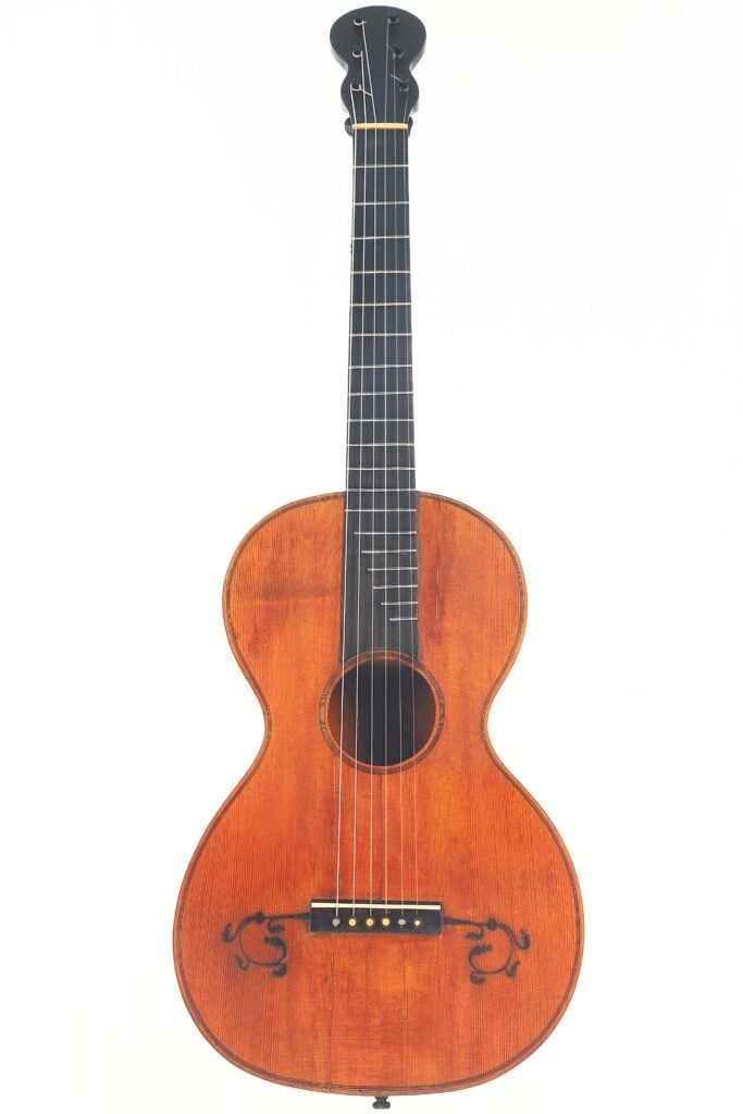 Die „Italienische Romantikgitarre 1860“ ist eine antike Akustikgitarre mit einem warmbraunen Holzkorpus, einem dunklen Griffbrett, sechs Saiten und schwarzen Verzierungen im Bereich des Stegs, fotografiert vor einem schlichten weißen Hintergrund. Die „Italienische Romantikgitarre 1860“ ist eine antike Akustikgitarre mit einem warmbraunen Holzkorpus, einem dunklen Griffbrett, sechs Saiten und schwarzen Verzierungen im Bereich des Stegs, fotografiert vor einem schlichten weißen Hintergrund.