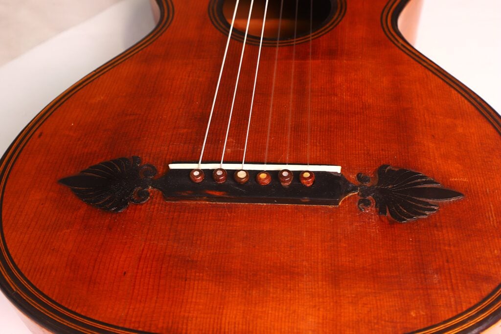 Nahaufnahme des Holzkörpers der „Romantikgitarre“ von Stephan Thumhart aus dem Jahr 1838, die sich durch eine satte rotbraune Lackierung, sechs am Steg befestigte Saiten und dekorative, geschnitzte Blattmotive auf beiden Seiten auszeichnet. Nahaufnahme des Holzkörpers der „Romantikgitarre“ von Stephan Thumhart aus dem Jahr 1838, die sich durch eine satte rotbraune Lackierung, sechs am Steg befestigte Saiten und dekorative, geschnitzte Blattmotive auf beiden Seiten auszeichnet.