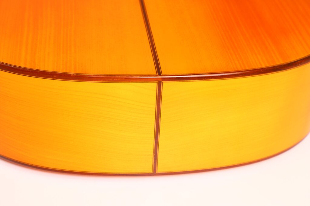 Nahaufnahme der Rückseite und der Seite der Juan Montero 1993 Blanca Flamenco-Gitarre. Man erkennt die glatte, polierte Oberfläche, die sichtbare Holzmaserung, das glänzende Finish und den präzisen Treffpunkt der Platten. Nahaufnahme der Rückseite und der Seite der Juan Montero 1993 Blanca Flamenco-Gitarre. Man erkennt die glatte, polierte Oberfläche, die sichtbare Holzmaserung, das glänzende Finish und den präzisen Treffpunkt der Platten.