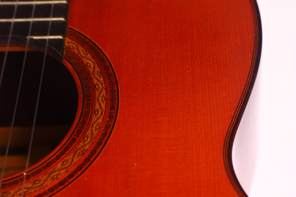 Nahaufnahme einer „Klassische Gitarre“ von Joachim Schneider aus dem Jahr 1977, bei der das Schallloch, die Zierrosette, die Saiten und der rotbraune Holzkorpus vor weißem Hintergrund hervorgehoben werden. Nahaufnahme einer „Klassische Gitarre“ von Joachim Schneider aus dem Jahr 1977, bei der das Schallloch, die Zierrosette, die Saiten und der rotbraune Holzkorpus vor weißem Hintergrund hervorgehoben werden.
