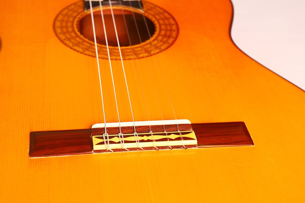 Nahaufnahme des Korpus einer Juan Montero 1993 Blanca Flamenco Gitarre mit den Saiten, dem Steg und einem Teil des Schalllochs auf einer warmen Holzoberfläche. Nahaufnahme des Korpus einer Juan Montero 1993 Blanca Flamenco Gitarre mit den Saiten, dem Steg und einem Teil des Schalllochs auf einer warmen Holzoberfläche.