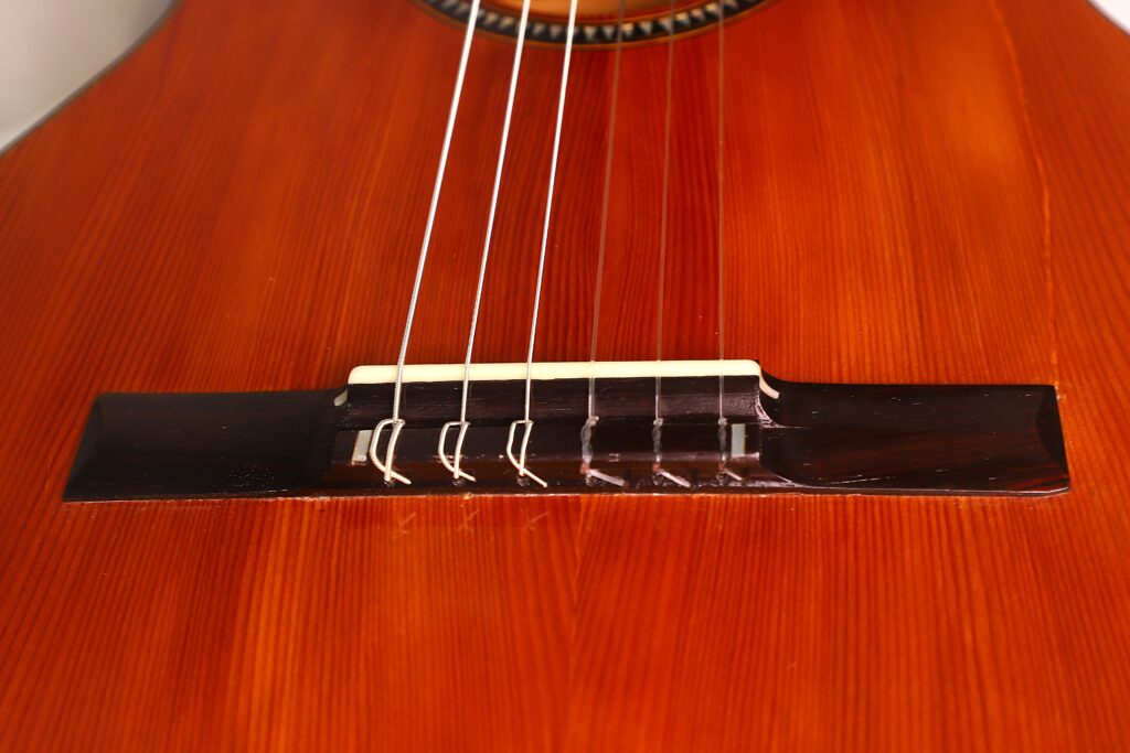 Nahaufnahme des Stegs und der Saiten der Andres Marin 1890 Klassische Gitarre auf einem polierten Holzkorpus, der die feine Holzmaserung und die detaillierte Handwerkskunst hervorhebt. Nahaufnahme des Stegs und der Saiten der Andres Marin 1890 Klassische Gitarre auf einem polierten Holzkorpus, der die feine Holzmaserung und die detaillierte Handwerkskunst hervorhebt.