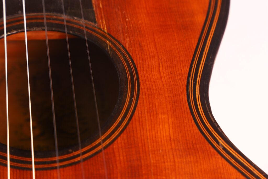 Nahaufnahme des Korpus der „Romantikgitarre“ von Stephan Thumhart aus dem Jahr 1838, die das Schallloch, die Saiten und die feine Holzmaserung in warmen Braun- und Orangetönen hervorhebt. Nahaufnahme des Korpus der „Romantikgitarre“ von Stephan Thumhart aus dem Jahr 1838, die das Schallloch, die Saiten und die feine Holzmaserung in warmen Braun- und Orangetönen hervorhebt.