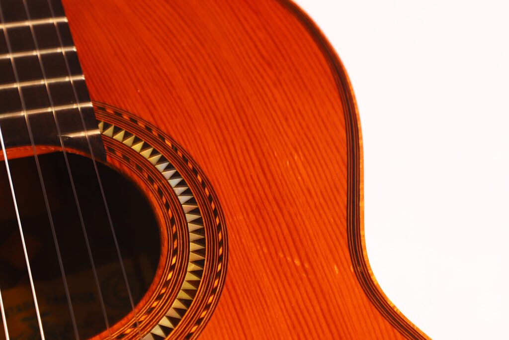 Nahaufnahme des Korpus der Andres Marin 1900 Klassische Gitarre. Hervorzuheben sind das Schallloch mit dekorativer Einlage, die sichtbaren Saiten und die reiche Holzmaserung vor weißem Hintergrund. Nahaufnahme des Korpus der Andres Marin 1900 Klassische Gitarre. Hervorzuheben sind das Schallloch mit dekorativer Einlage, die sichtbaren Saiten und die reiche Holzmaserung vor weißem Hintergrund.