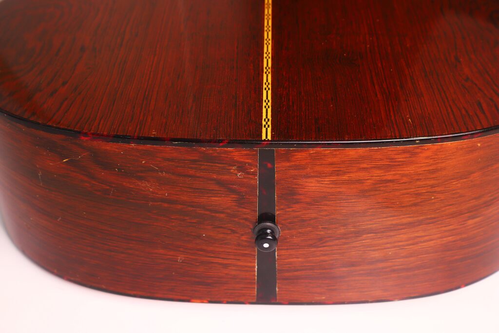 Nahaufnahme der Rückseite und der Seite einer Martin D-21 Akustikgitarre aus dem Jahr 1965 mit einer dekorativen Einlage und einem Gurtknopf am unteren Rand. Nahaufnahme der Rückseite und der Seite einer Martin D-21 Akustikgitarre aus dem Jahr 1965 mit einer dekorativen Einlage und einem Gurtknopf am unteren Rand.