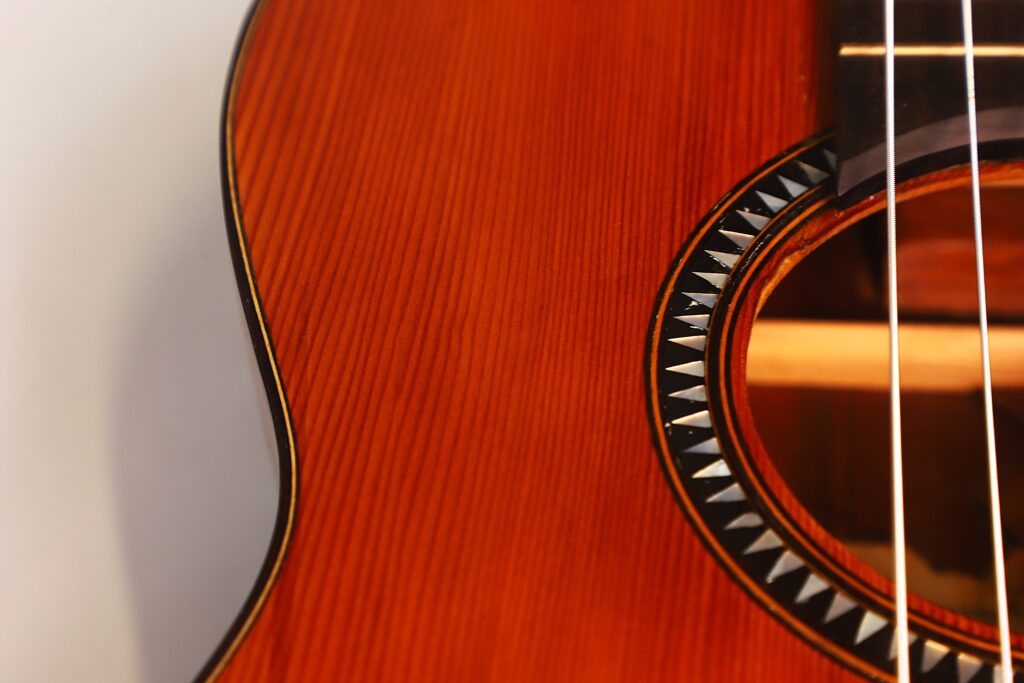 Nahaufnahme des Korpus der Andres Marin 1890 Klassische Gitarre. Hervorzuheben sind die hölzerne Textur, ein Teil des Schalllochs mit dekorativem Muster und drei sichtbare Gitarrensaiten. Nahaufnahme des Korpus der Andres Marin 1890 Klassische Gitarre. Hervorzuheben sind die hölzerne Textur, ein Teil des Schalllochs mit dekorativem Muster und drei sichtbare Gitarrensaiten.