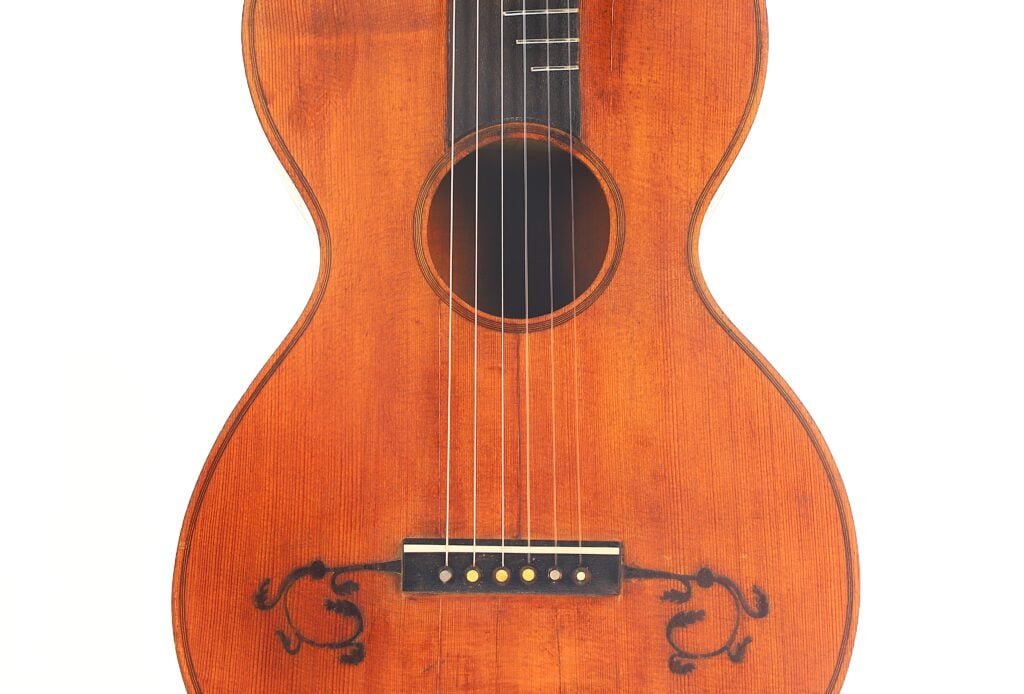 Nahaufnahme der „Italienischen Romantikgitarre 1860“ mit warmer brauner Lackierung, sechs Saiten, rundem Schallloch und schwarzen Verzierungen in der Nähe des Stegs vor weißem Hintergrund. Nahaufnahme der „Italienischen Romantikgitarre 1860“ mit warmer brauner Lackierung, sechs Saiten, rundem Schallloch und schwarzen Verzierungen in der Nähe des Stegs vor weißem Hintergrund.