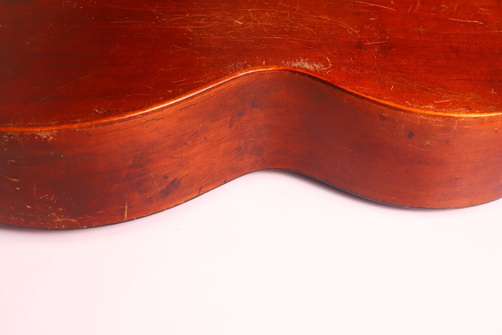 Nahaufnahme des hölzernen Korpus einer Braun & Hauser 1905 Klassische Gitarre. Hervorzuheben sind die gewölbte Kante und die sichtbaren Kratzer und Abnutzungserscheinungen auf der Oberfläche, die sich vor einem schlichten hellen Hintergrund befinden. Nahaufnahme des hölzernen Korpus einer Braun & Hauser 1905 Klassische Gitarre. Hervorzuheben sind die gewölbte Kante und die sichtbaren Kratzer und Abnutzungserscheinungen auf der Oberfläche, die sich vor einem schlichten hellen Hintergrund befinden.