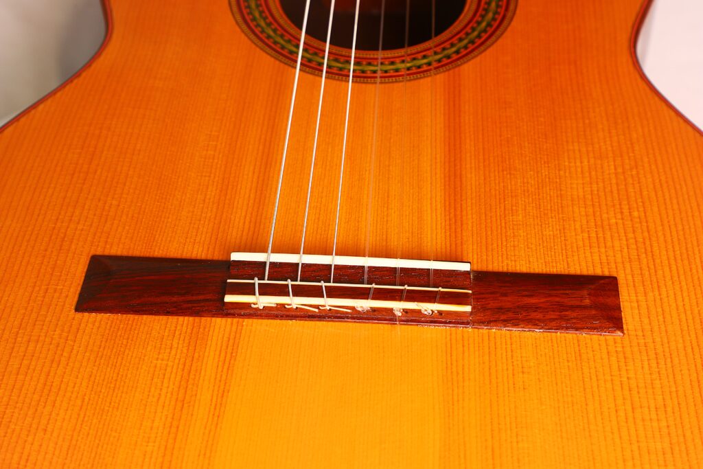Nahaufnahme des Korpus der klassischen Gitarre „Carl Hermann Schäfer – Nicolaus Wollf 2012“, mit Blick auf den Steg aus Holz, die Saiten und das Schallloch mit dekorativer Rosette. Mit warmer orange-brauner Lackierung und sichtbarer Holzmaserung. Nahaufnahme des Korpus der klassischen Gitarre „Carl Hermann Schäfer – Nicolaus Wollf 2012“, mit Blick auf den Steg aus Holz, die Saiten und das Schallloch mit dekorativer Rosette. Mit warmer orange-brauner Lackierung und sichtbarer Holzmaserung.