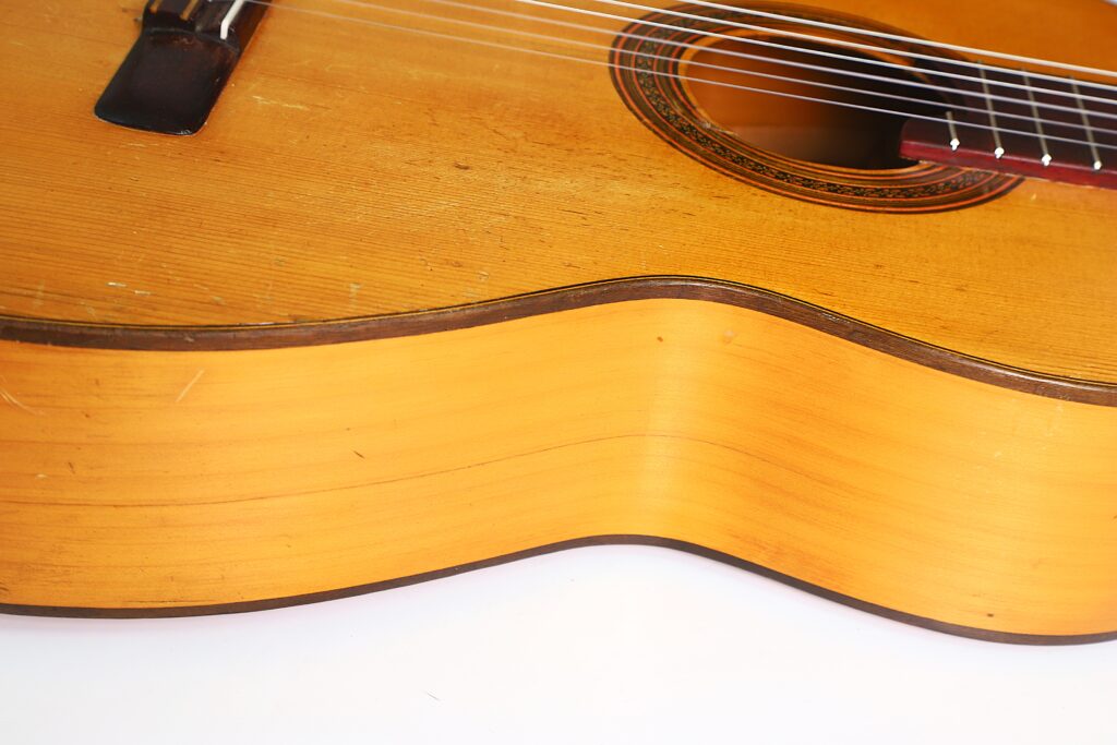 Nahaufnahme des Korpus der „Jose Ramirez I 1921 Klassische Gitarre“, die die Holzmaserung, den Steg, das Schallloch und die Saiten vor einem weißen Hintergrund hervorhebt. Nahaufnahme des Korpus der „Jose Ramirez I 1921 Klassische Gitarre“, die die Holzmaserung, den Steg, das Schallloch und die Saiten vor einem weißen Hintergrund hervorhebt.