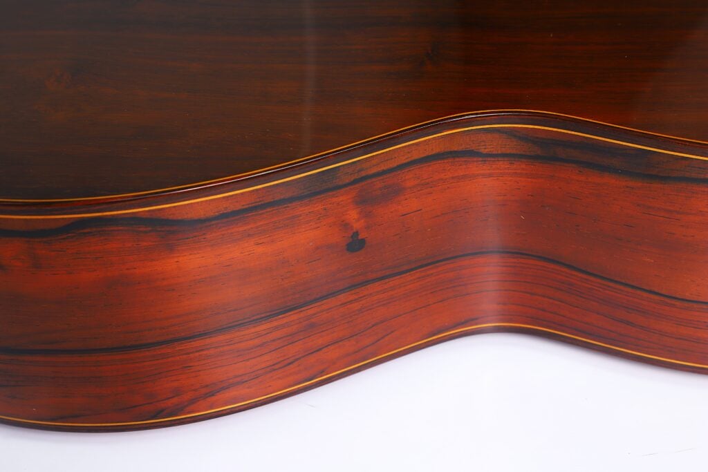 Nahaufnahme der Seite einer „Doble Tapa Klassische Gitarre“ von Manuel Contreras aus dem Jahr 1981, die sich durch eine satte rotbraune Holzmaserung mit dunklen Streifen und eine glatte, glänzende Oberfläche auszeichnet. Die Rundungen des Gitarrenkorpus bilden ein sanftes Wellenmuster. Nahaufnahme der Seite einer „Doble Tapa Klassische Gitarre“ von Manuel Contreras aus dem Jahr 1981, die sich durch eine satte rotbraune Holzmaserung mit dunklen Streifen und eine glatte, glänzende Oberfläche auszeichnet. Die Rundungen des Gitarrenkorpus bilden ein sanftes Wellenmuster.