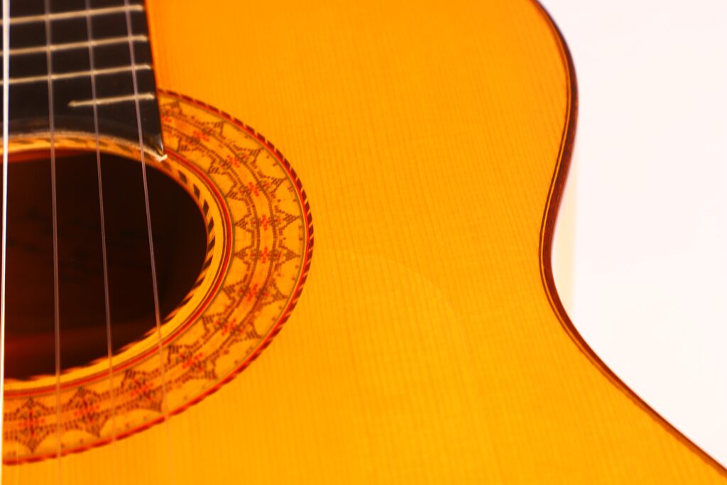 Nahaufnahme einer Juan Montero 1993 Blanca Flamenco-Gitarre. Hervorzuheben sind die detaillierte Holzmaserung, die verzierte Rosette um das Schallloch sowie Teile der Saiten und des Griffbretts vor einem weißen Hintergrund. Nahaufnahme einer Juan Montero 1993 Blanca Flamenco-Gitarre. Hervorzuheben sind die detaillierte Holzmaserung, die verzierte Rosette um das Schallloch sowie Teile der Saiten und des Griffbretts vor einem weißen Hintergrund.