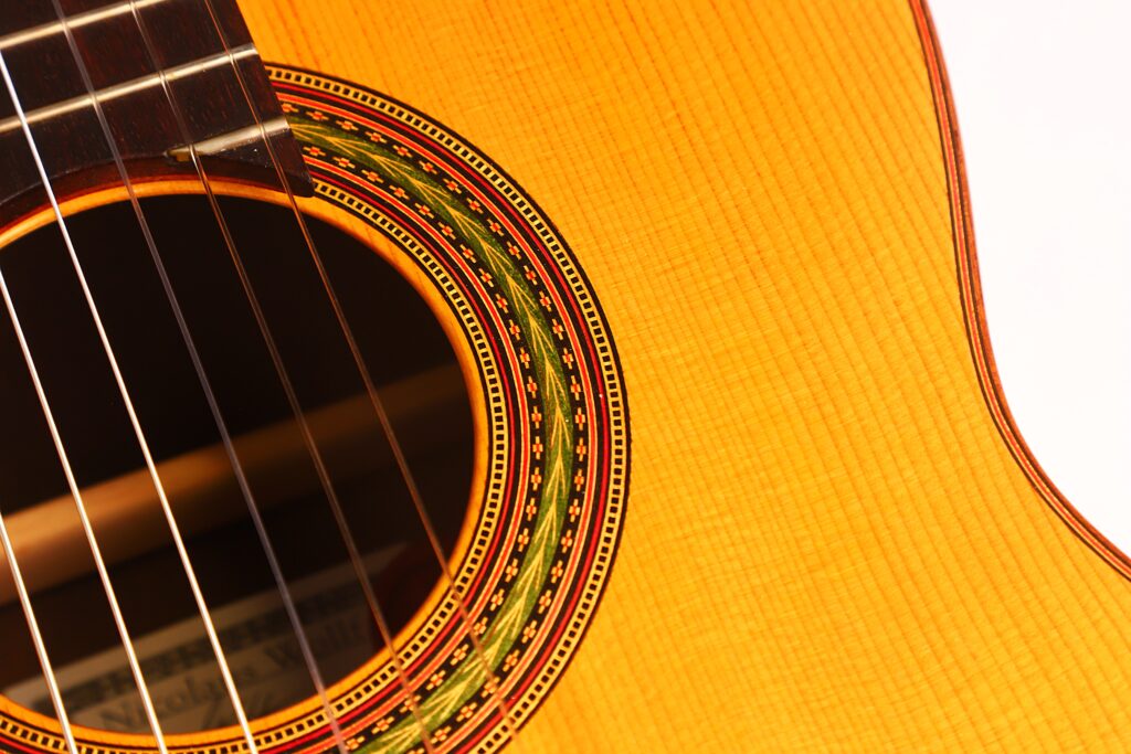 Nahaufnahme des Schalllochs und der Saiten der „Carl Hermann Schäfer – Nicolaus Wollf 2012 Klassische Gitarre“, mit einer detailreichen Rosette und einem Korpus mit sichtbarer Holzmaserung, wobei die Beleuchtung die warme, goldbraune Farbe hervorhebt. Nahaufnahme des Schalllochs und der Saiten der „Carl Hermann Schäfer – Nicolaus Wollf 2012 Klassische Gitarre“, mit einer detailreichen Rosette und einem Korpus mit sichtbarer Holzmaserung, wobei die Beleuchtung die warme, goldbraune Farbe hervorhebt.