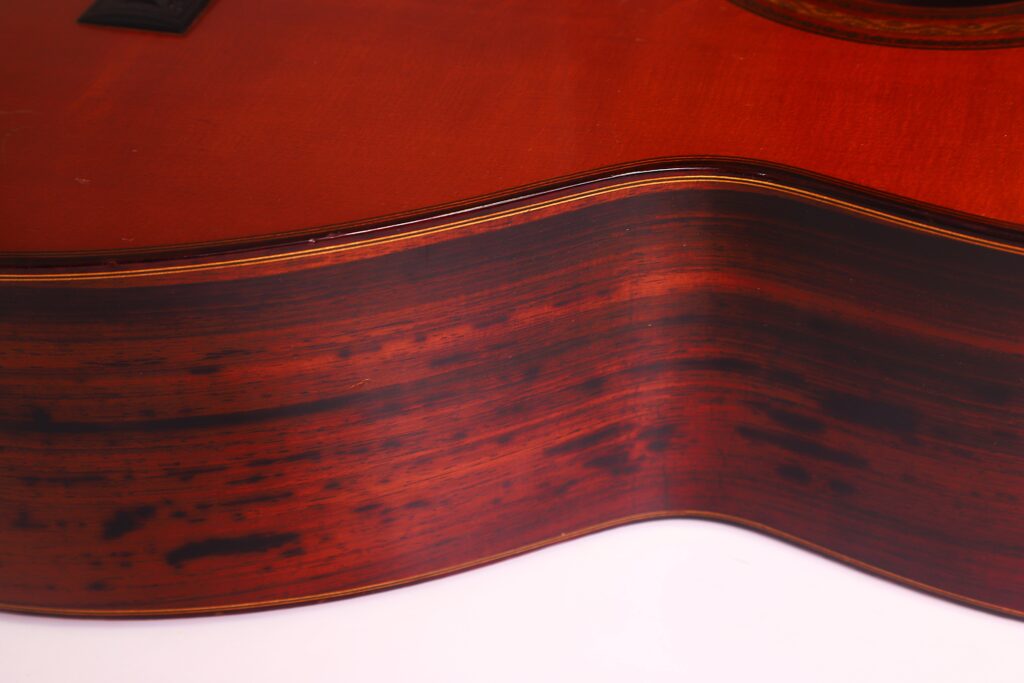 Nahaufnahme der Seite der „Joachim Schneider 1977 Klassische Gitarre“, die die Holzmaserung und die rotbraune Lackierung mit dunklen Streifen zeigt und die elegante Korpuswölbung des Instruments vor einem weißen Hintergrund hervorhebt. Nahaufnahme der Seite der „Joachim Schneider 1977 Klassische Gitarre“, die die Holzmaserung und die rotbraune Lackierung mit dunklen Streifen zeigt und die elegante Korpuswölbung des Instruments vor einem weißen Hintergrund hervorhebt.