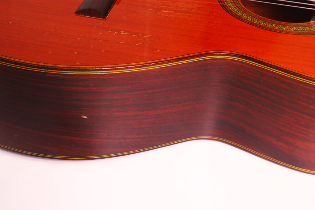 Nahaufnahme der Seite und des unteren Teils des Korpus einer „Juan Estruch Chet Atkins Yellow Label 1976 Klassische Gitarre“, die die Holzmaserung, leichte Kratzer und eine kleine Delle auf der dunkel polierten Oberfläche vor einem schlichten weißen Hintergrund zeigt. Nahaufnahme der Seite und des unteren Teils des Korpus einer „Juan Estruch Chet Atkins Yellow Label 1976 Klassische Gitarre“, die die Holzmaserung, leichte Kratzer und eine kleine Delle auf der dunkel polierten Oberfläche vor einem schlichten weißen Hintergrund zeigt.