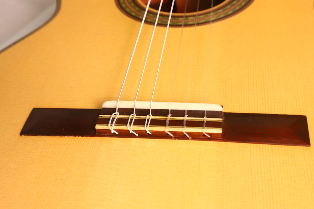 Nahaufnahme von Steg und Saiten der Antonio Aparicio Mod. Master Klassische Gitarre, mit Holzmaserung und Nylonsaiten auf dem dunklen Holzsteg. Nahaufnahme von Steg und Saiten der Antonio Aparicio Mod. Master Klassische Gitarre, mit Holzmaserung und Nylonsaiten auf dem dunklen Holzsteg.