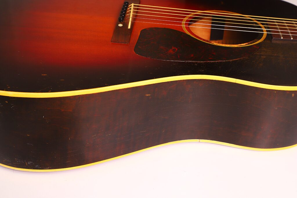 Nahaufnahme der Gibson J-45 Banner 1944 Akustik-Gitarre mit Sunburst-Lackierung, die die Saiten, den Steg und das Schlagbrett vor einem schlichten weißen Hintergrund zeigt. Nahaufnahme der Gibson J-45 Banner 1944 Akustik-Gitarre mit Sunburst-Lackierung, die die Saiten, den Steg und das Schlagbrett vor einem schlichten weißen Hintergrund zeigt.