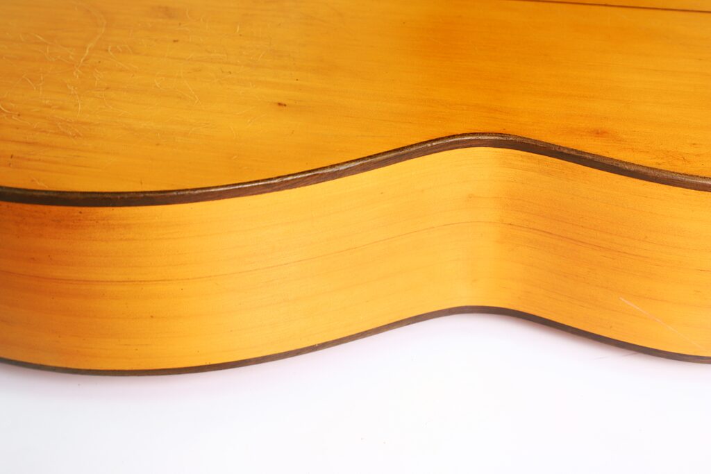 Nahaufnahme der Seite und des Randes einer „Jose Ramirez I 1921“ Klassischen Gitarre mit heller Holzmaserung, dunkler Einfassung und ihrer elegant geschwungenen Form vor weißem Hintergrund. Nahaufnahme der Seite und des Randes einer „Jose Ramirez I 1921“ Klassischen Gitarre mit heller Holzmaserung, dunkler Einfassung und ihrer elegant geschwungenen Form vor weißem Hintergrund.