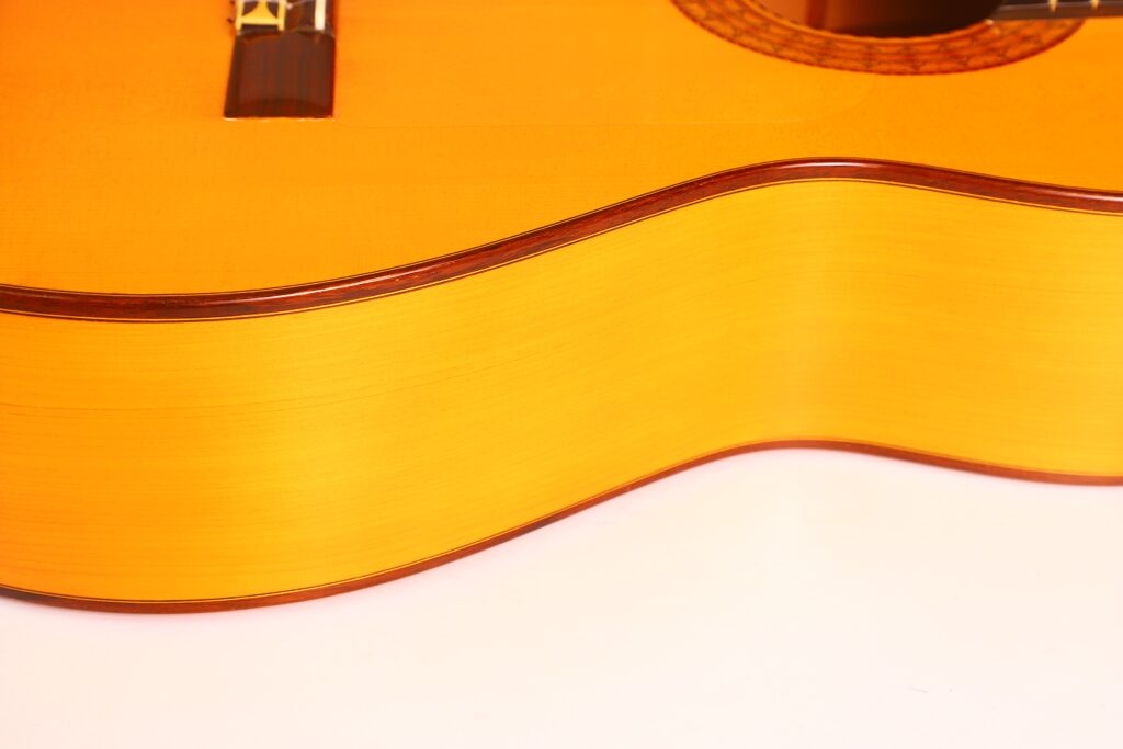 Nahaufnahme der Seite und des Korpus einer Juan Montero 1993 Blanca Flamenco-Gitarre. Hervorzuheben sind die glatte Holzoberfläche, die elegante geschwungene Form und die leicht gelblich-braune Farbe vor weißem Hintergrund. Nahaufnahme der Seite und des Korpus einer Juan Montero 1993 Blanca Flamenco-Gitarre. Hervorzuheben sind die glatte Holzoberfläche, die elegante geschwungene Form und die leicht gelblich-braune Farbe vor weißem Hintergrund.