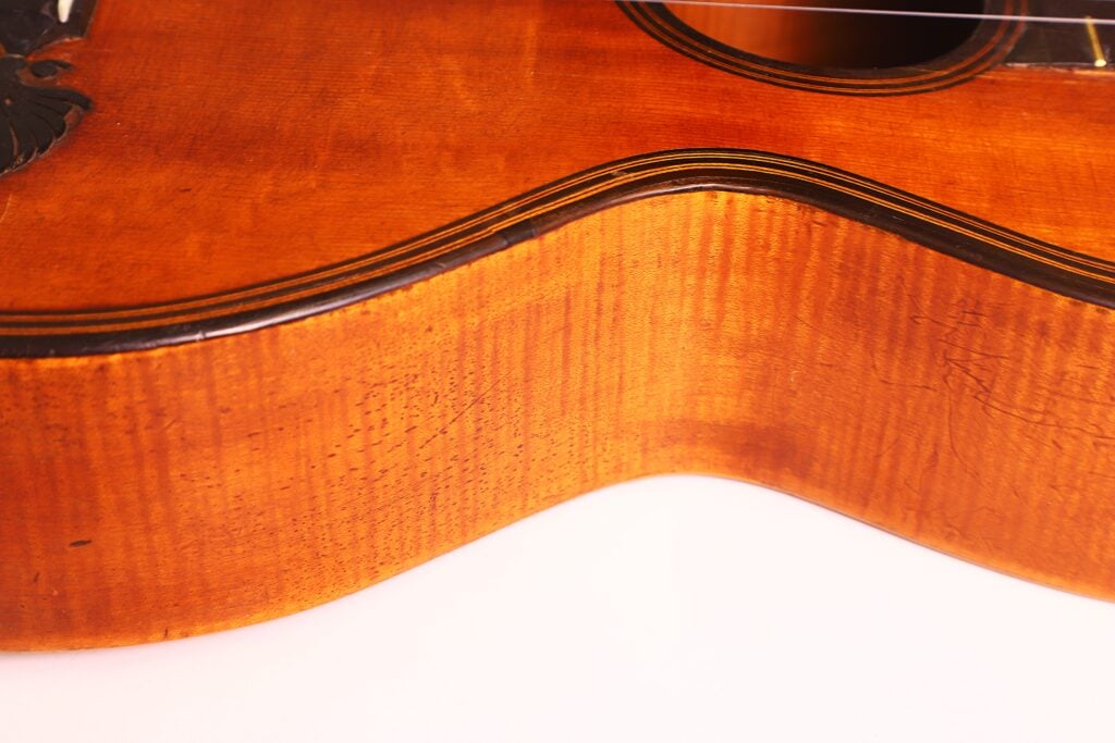 Nahaufnahme der Zargen und des Korpus der „Romantikgitarre“ von Stephan Thumhart aus dem Jahr 1838, die vor einem schlichten weißen Hintergrund die Maserung des Holzes, die natürliche Farbe, feine Oberflächenrisse und das teilweise sichtbare Schallloch hervorhebt. Nahaufnahme der Zargen und des Korpus der „Romantikgitarre“ von Stephan Thumhart aus dem Jahr 1838, die vor einem schlichten weißen Hintergrund die Maserung des Holzes, die natürliche Farbe, feine Oberflächenrisse und das teilweise sichtbare Schallloch hervorhebt.