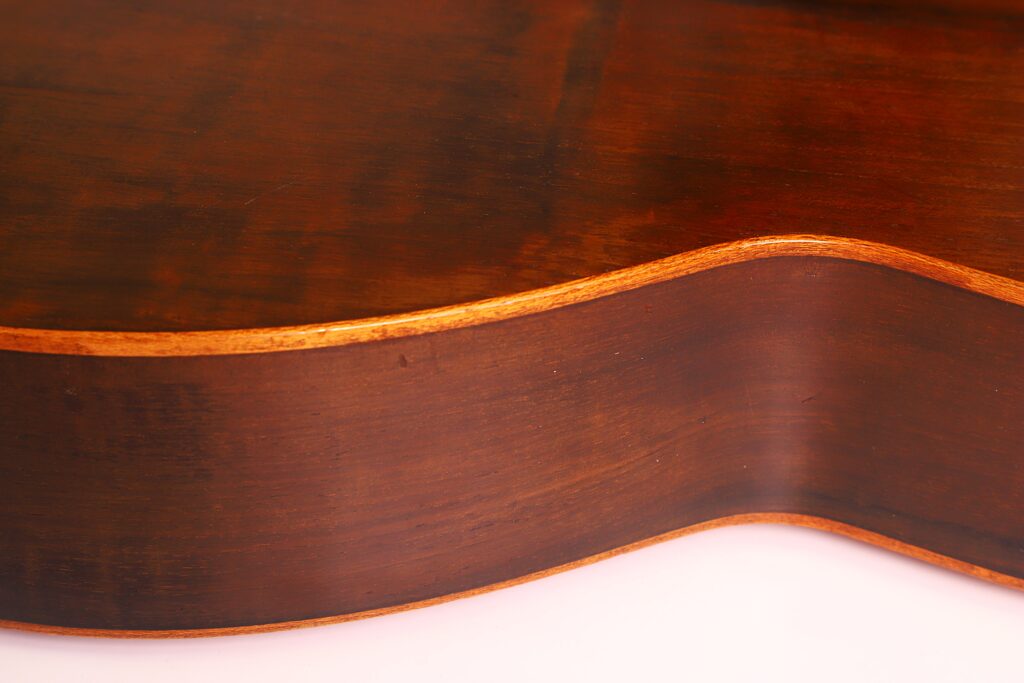 Nahaufnahme der gewölbten Seite der Andres Marin 1900 Klassische Gitarre, wobei die glatte, polierte braune Holzmaserung und die hellere Kante vor einem hellen Hintergrund hervorgehoben werden. Nahaufnahme der gewölbten Seite der Andres Marin 1900 Klassische Gitarre, wobei die glatte, polierte braune Holzmaserung und die hellere Kante vor einem hellen Hintergrund hervorgehoben werden.