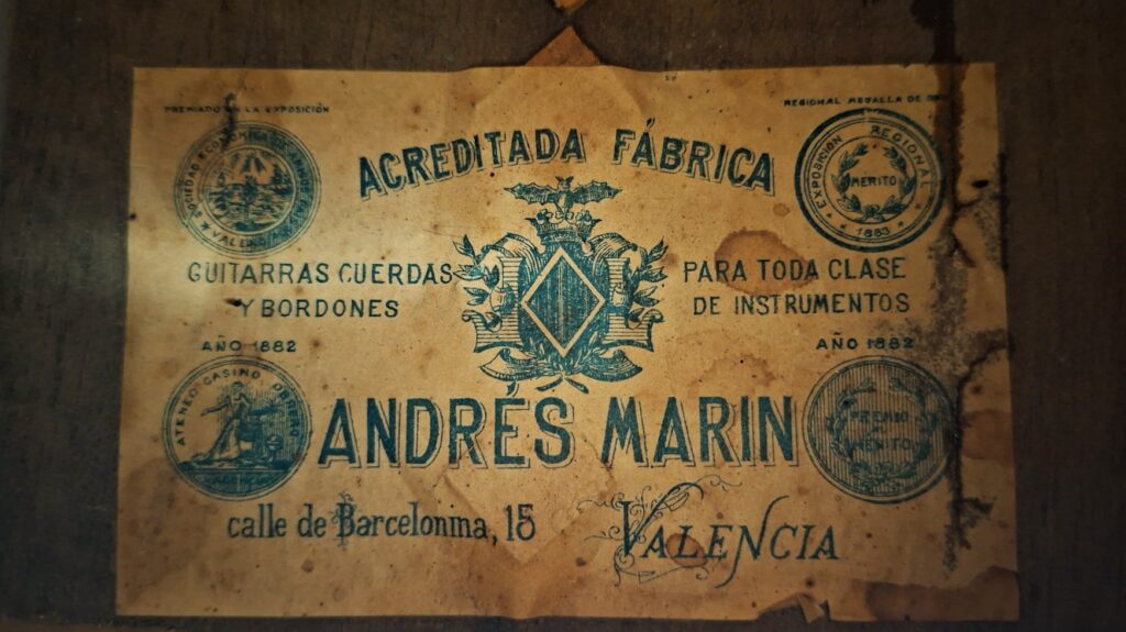 Ein Vintage-Etikett für die Andres Marin 1900 Klassische Gitarre aus Valencia, Spanien, zeigt verzierten blauen Text, dekorative Embleme und Stempel und listet Guitarras Cuerdas y Bordones mit der Adresse Calle de Barcelona, 15. Ein Vintage-Etikett für die Andres Marin 1900 Klassische Gitarre aus Valencia, Spanien, zeigt verzierten blauen Text, dekorative Embleme und Stempel und listet Guitarras Cuerdas y Bordones mit der Adresse Calle de Barcelona, 15.