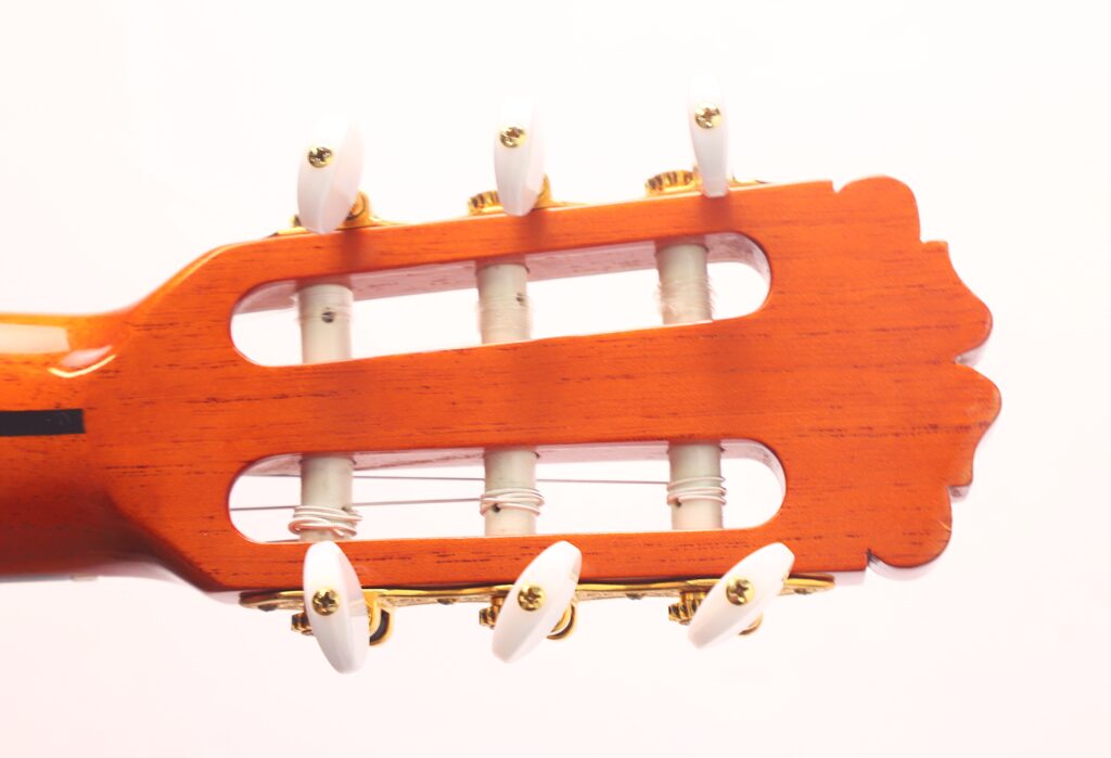 Nahaufnahme der Kopfplatte der Juan Miguel Gonzalez 2006 Klassische Gitarre mit weißen Stimmwirbeln, goldener Hardware und Nylonsaiten vor hellem Hintergrund. Nahaufnahme der Kopfplatte der Juan Miguel Gonzalez 2006 Klassische Gitarre mit weißen Stimmwirbeln, goldener Hardware und Nylonsaiten vor hellem Hintergrund.