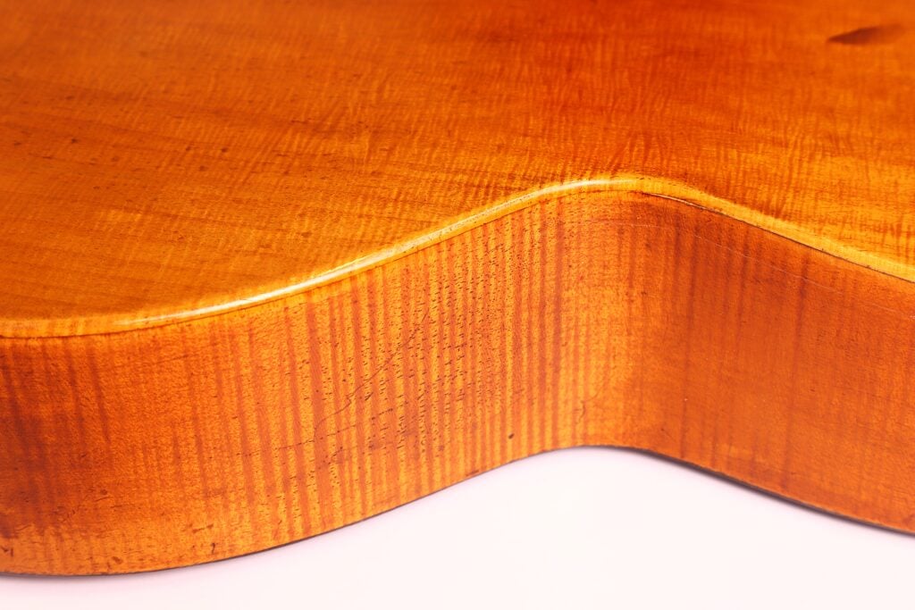 Nahaufnahme der Seite und der geschwungenen Kante der „Romantikgitarre“ von Stephan Thumhart aus dem Jahr 1838, die die glatte, polierte, goldbraune Maserung und die feine Struktur des Holzes zeigt. Nahaufnahme der Seite und der geschwungenen Kante der „Romantikgitarre“ von Stephan Thumhart aus dem Jahr 1838, die die glatte, polierte, goldbraune Maserung und die feine Struktur des Holzes zeigt.