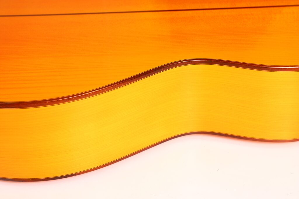 Nahaufnahme der Seite einer Juan Montero 1993 Blanca Flamenco-Gitarre mit ihrem glatten, hellen, gewölbten Korpus mit glänzender Oberfläche und dunklen Rändern vor einem schlichten Hintergrund. Nahaufnahme der Seite einer Juan Montero 1993 Blanca Flamenco-Gitarre mit ihrem glatten, hellen, gewölbten Korpus mit glänzender Oberfläche und dunklen Rändern vor einem schlichten Hintergrund.