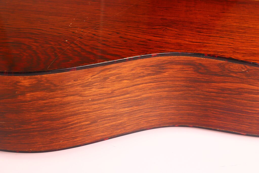 Nahaufnahme der Seite und der Rückseite der Martin D-21 1965 Akustik Gitarre, die die glatte, glänzende Oberfläche und die reiche rötlich-braune Holzmaserung mit Lichtreflexionen auf der gewölbten Oberfläche zeigt. Nahaufnahme der Seite und der Rückseite der Martin D-21 1965 Akustik Gitarre, die die glatte, glänzende Oberfläche und die reiche rötlich-braune Holzmaserung mit Lichtreflexionen auf der gewölbten Oberfläche zeigt.