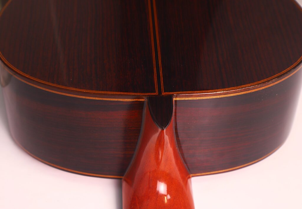 Nahaufnahme des Bodens und des Halses der Juan Miguel Gonzalez 2006 Klassische Gitarre mit poliertem, dunklem Holz, einer zentralen Naht und feinem Zierband. Nahaufnahme des Bodens und des Halses der Juan Miguel Gonzalez 2006 Klassische Gitarre mit poliertem, dunklem Holz, einer zentralen Naht und feinem Zierband.