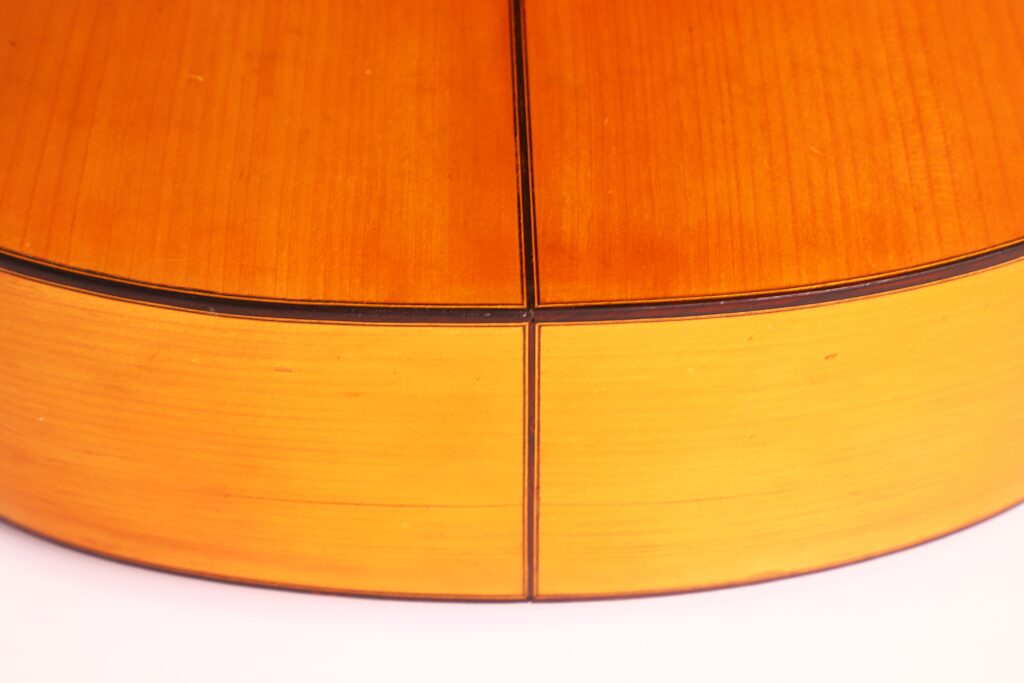 Nahaufnahme des Bodens und der Seite einer Flamenco-Gitarre von Jose Ramirez aus dem Jahr 1955. Das Holz ist hellbraun mit sichtbarer Maserung und dunklen Einlegearbeiten, die ein Kreuzmuster bilden. Nahaufnahme des Bodens und der Seite einer Flamenco-Gitarre von Jose Ramirez aus dem Jahr 1955. Das Holz ist hellbraun mit sichtbarer Maserung und dunklen Einlegearbeiten, die ein Kreuzmuster bilden.
