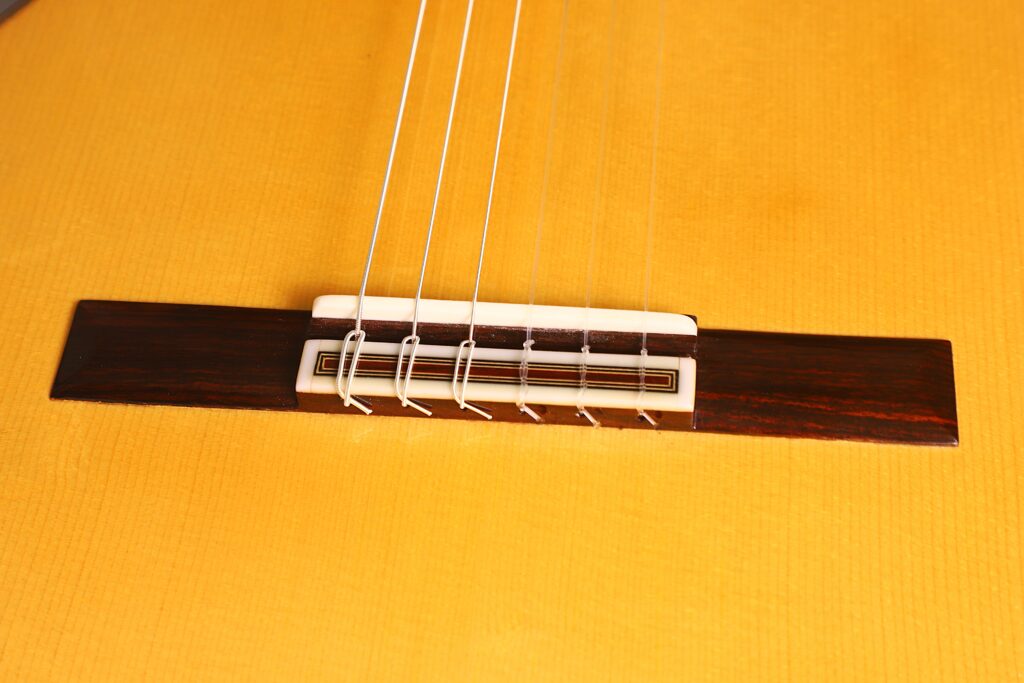 Nahaufnahme des Stegs und der Saiten einer Vicente Carrillo 1998 Torres Klassische Gitarre, wobei die Holzmaserung und die Details der Saitenbefestigung hervorgehoben werden. Nahaufnahme des Stegs und der Saiten einer Vicente Carrillo 1998 Torres Klassische Gitarre, wobei die Holzmaserung und die Details der Saitenbefestigung hervorgehoben werden.