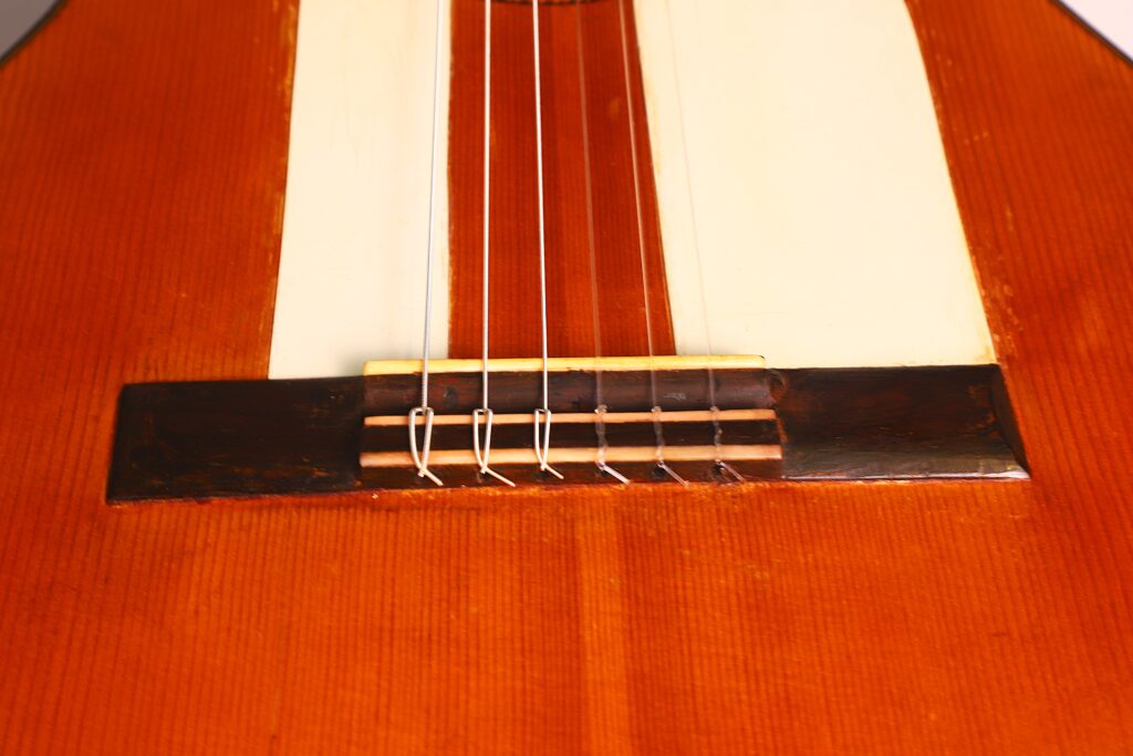 Nahaufnahme des Stegs und der Saiten einer Jose Ramirez 1955 Flamenco-Gitarre, die den hölzernen Korpus, den Sattel und das glänzende Finish hervorhebt. Nahaufnahme des Stegs und der Saiten einer Jose Ramirez 1955 Flamenco-Gitarre, die den hölzernen Korpus, den Sattel und das glänzende Finish hervorhebt.