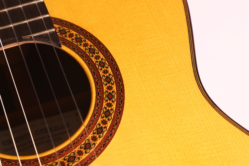 Nahaufnahme des Schalllochs der Vicente Carrillo 1998 Torres Klassische Gitarre, wobei die detaillierte Rosette, die Saiten und die Korpuskante vor einem schlichten hellen Hintergrund hervorgehoben werden. Nahaufnahme des Schalllochs der Vicente Carrillo 1998 Torres Klassische Gitarre, wobei die detaillierte Rosette, die Saiten und die Korpuskante vor einem schlichten hellen Hintergrund hervorgehoben werden.