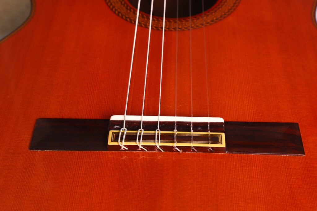 Nahaufnahme des Stegs der Juan Miguel Gonzalez 2006 Klassische Gitarre, wobei die am Holzsteg befestigten Saiten auf dem rötlich-braunen Korpus mit sichtbarer feiner Holzmaserung und dekorativer Einlage hervorgehoben werden. Nahaufnahme des Stegs der Juan Miguel Gonzalez 2006 Klassische Gitarre, wobei die am Holzsteg befestigten Saiten auf dem rötlich-braunen Korpus mit sichtbarer feiner Holzmaserung und dekorativer Einlage hervorgehoben werden.