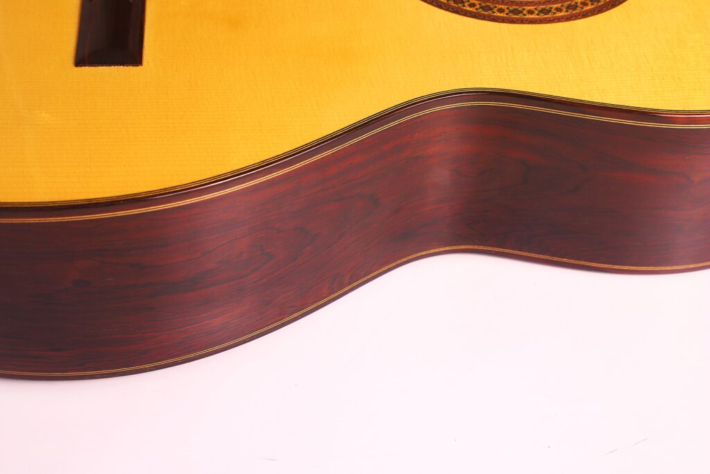 Nahaufnahme der Seite und der Oberseite einer Vicente Carrillo 1998 Torres Klassische Gitarre, wobei die Holzmaserung und die glatte, polierte Oberfläche vor einem weißen Hintergrund hervorgehoben werden. Nahaufnahme der Seite und der Oberseite einer Vicente Carrillo 1998 Torres Klassische Gitarre, wobei die Holzmaserung und die glatte, polierte Oberfläche vor einem weißen Hintergrund hervorgehoben werden.
