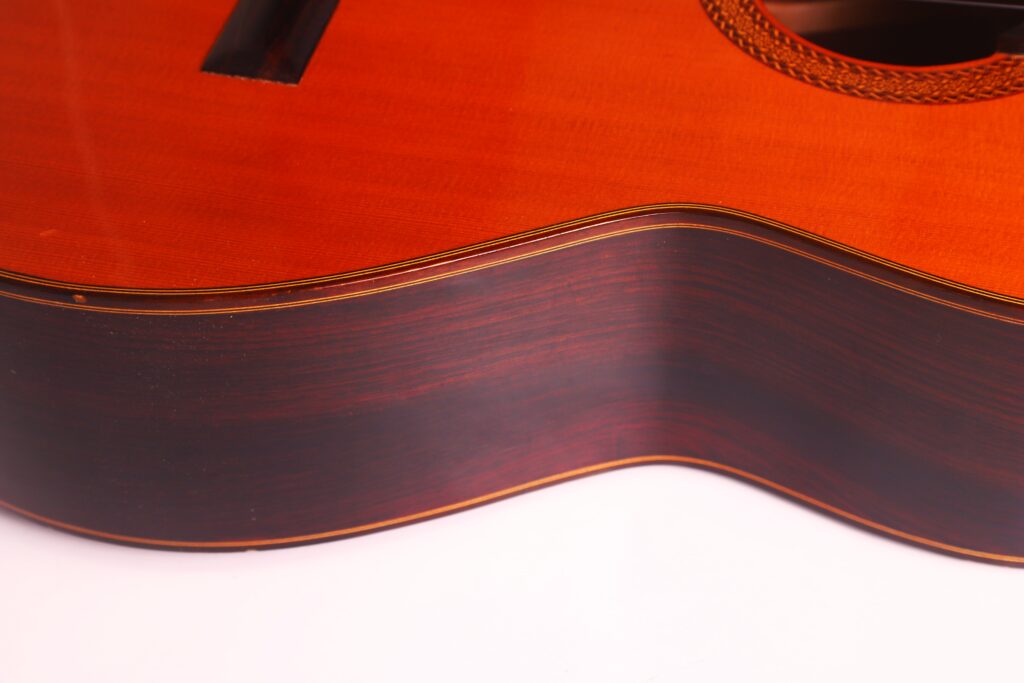 Nahaufnahme der Seite und des Korpus der Juan Miguel Gonzalez 2006 Klassische Gitarre. Hervorzuheben sind die Holzmaserung, die glänzende Oberfläche und die dekorativen Einfassungen vor einem weißen Hintergrund. Nahaufnahme der Seite und des Korpus der Juan Miguel Gonzalez 2006 Klassische Gitarre. Hervorzuheben sind die Holzmaserung, die glänzende Oberfläche und die dekorativen Einfassungen vor einem weißen Hintergrund.