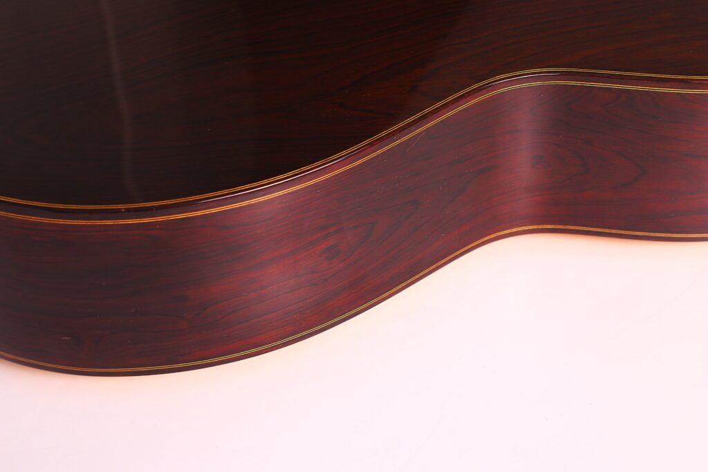 Nahaufnahme der polierten Holzseite einer Vicente Carrillo 1998 Torres Klassische Gitarre, wobei die glatte, dunkle Holzmaserung und die hellen Verzierungen vor einem hellen Hintergrund hervorgehoben werden. Nahaufnahme der polierten Holzseite einer Vicente Carrillo 1998 Torres Klassische Gitarre, wobei die glatte, dunkle Holzmaserung und die hellen Verzierungen vor einem hellen Hintergrund hervorgehoben werden.