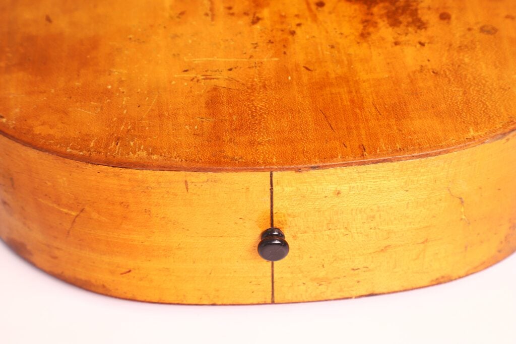 Eine Nahaufnahme der Unterkante der „Italienische Romantikgitarre 1860“ zeigt ihre abgerundete, glänzende, abgenutzte gelb-orangefarbene Lackierung und einen einzelnen schwarzen Gurtknopf in der Mitte. Eine Nahaufnahme der Unterkante der „Italienische Romantikgitarre 1860“ zeigt ihre abgerundete, glänzende, abgenutzte gelb-orangefarbene Lackierung und einen einzelnen schwarzen Gurtknopf in der Mitte.