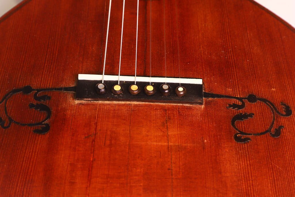 Nahaufnahme der „Italienischen Romantikgitarre 1860“ mit einem rotbraunen Holzkorpus, sechs Saiten über dem Steg und dekorativen schwarzen Wirbelmotiven, die den Steg flankieren. Nahaufnahme der „Italienischen Romantikgitarre 1860“ mit einem rotbraunen Holzkorpus, sechs Saiten über dem Steg und dekorativen schwarzen Wirbelmotiven, die den Steg flankieren.