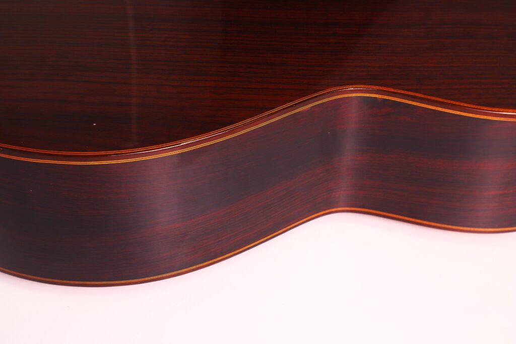 Nahaufnahme des glänzenden, dunkelbraunen Holzkorpus und der gewölbten Seite der Juan Miguel Gonzalez 2006 Klassische Gitarre, die eine feine Holzmaserung und eine dünne Zierleiste entlang der Kante zeigt. Nahaufnahme des glänzenden, dunkelbraunen Holzkorpus und der gewölbten Seite der Juan Miguel Gonzalez 2006 Klassische Gitarre, die eine feine Holzmaserung und eine dünne Zierleiste entlang der Kante zeigt.